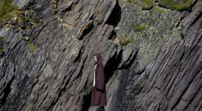Person in a brown cloak stands on a cliff, reminiscent of Slea Head Drive near Dingle.