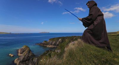 Person in a brown cloak with blue lightsaber stands on a grassy cliff on the Slea Head drive tour.